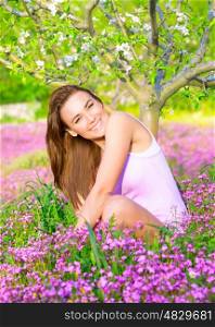 Cheerful smiling female spending time on backyard, enjoying apple tree blossom, sitting on pink floral glade, spring season concept
