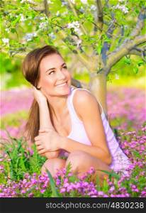 Cheerful smiling female spending time on backyard, looking on side, enjoying apple tree blossom, sitting on pink floral glade, spring season concept