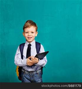 Cheerful smiling little boy with big backpack holding digital tablet against blue background. Looking at camera. School concept. Back to School