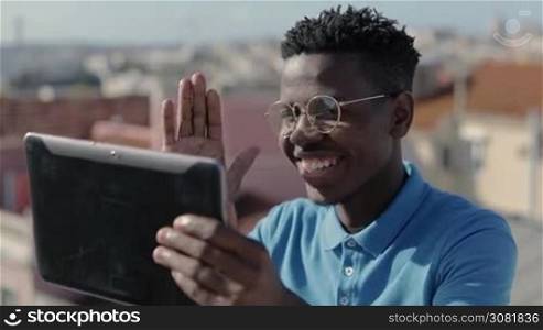 Cheerful smiling man greetings in video chat on digital tablet. African American young man shares old city roofs landscape with video chat. Travel impressions concept