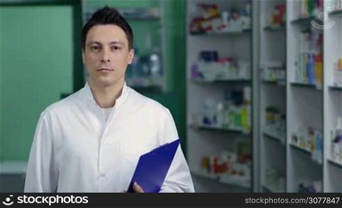 Cheerful smiling pharmacist chemist man with clipboard in hand standing in pharmacy drugstore. Positive male druggist in white coat posing and looking at the camera with toohy smile over drugstore background. Health care and pharmacology concept.