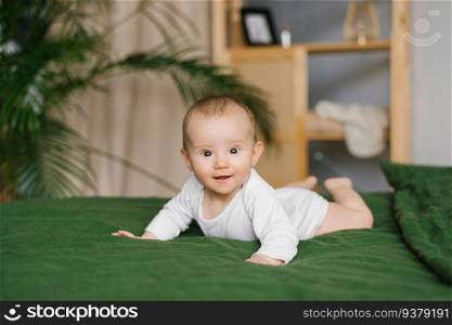 Cheerful smiling six-month-old baby in a bodysuit lies on his stomach on a cozy bed