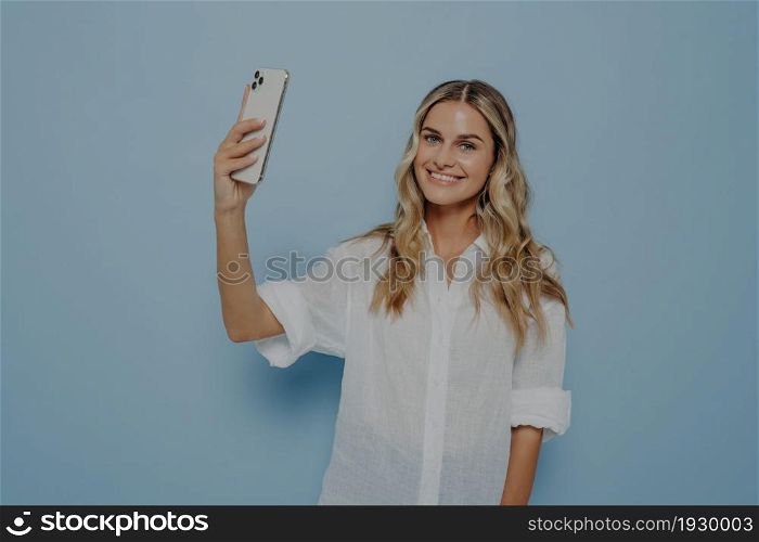 Cheerful smiling young woman with wavy long blond hair making photo on smartphone, happy female posing for selfie or photo shoot for blog, indoor studio image isolated on blue background. Cheerful smiling young woman with wavy long blond hair making photo on smartphone