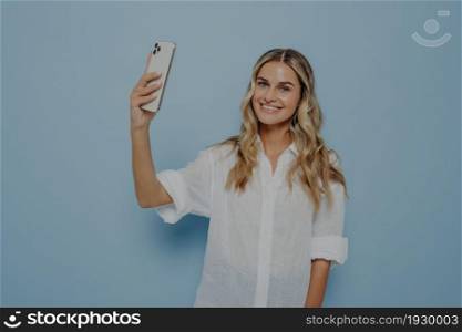 Cheerful smiling young woman with wavy long blond hair making photo on smartphone, happy female posing for selfie or photo shoot for blog, indoor studio image isolated on blue background. Cheerful smiling young woman with wavy long blond hair making photo on smartphone