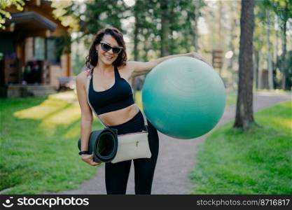 Cheerful sporty young woman dressed in active wear holds big fitness ball, wears sunglasses, carries karemat, has good mood, prepares for fitness training outdoor, has regular sport exercises