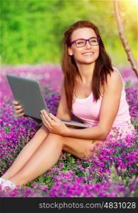 Cheerful student girl in the park, sitting on flower meadow and doing homework on laptop, studying in high school, back to school