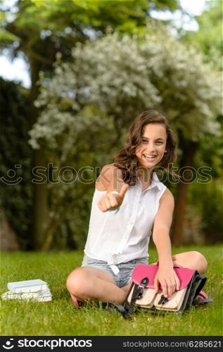 Cheerful student girl sitting on grass shows thumb up summer