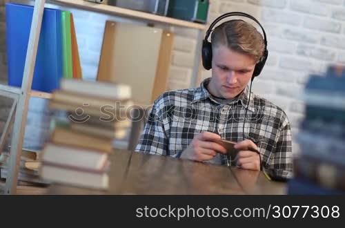 Cheerful student in library chatting on mobile phone while listening to music with headphones. Handsome teenager sitting at the table surrounded by many books and typing text message on smartphone while studying.