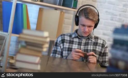 Cheerful student in library chatting on mobile phone while listening to music with headphones. Handsome teenager sitting at the table surrounded by many books and typing text message on smartphone while studying.