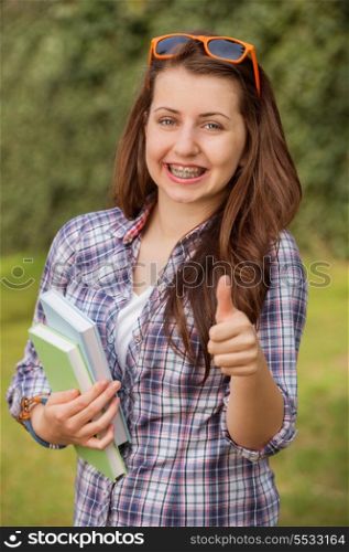 Cheerful student with braces holding books showing thumb up outside