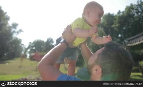 Cheerful stylish father with beard lifting cute baby boy up high in the air while spending and enjoying time together in park. Young dad expressing his love and tenderness, kissing his adorable infant child and having fun while raising child up.