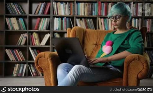 Cheerful successful freelancer girl in eyeglasses working on her laptop while sitting in comfortable chair at home over bookshelves background. Smiling intelligent hipster freelancer connecting to internet via computer and working on new project.