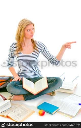 Cheerful teen girl sitting on floor among schoolbooks and pointing in corner isolated on white &#xA;