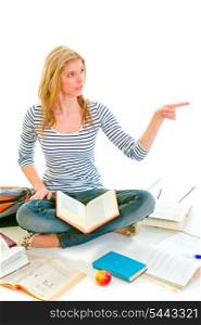Cheerful teen girl sitting on floor among schoolbooks and pointing in corner isolated on white &#xA;