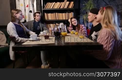 Cheerful teenagers meeting in cafe after college. Group of young friends having great time together at local coffee shop. Smiling people relaxing, chatting and discussing latest gossips while sitting at restaurant table served with coffee and juice.