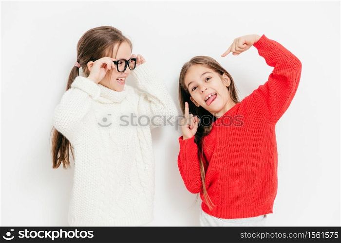 Cheerful two sisters have fun together, have funny expressions. Adorable child in eyewear and white warm sweater looks at her best female friend who makes grimace, isolated over studio background
