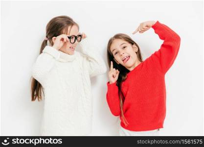Cheerful two sisters have fun together, have funny expressions. Adorable child in eyewear and white warm sweater looks at her best female friend who makes grimace, isolated over studio background