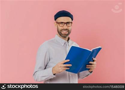Cheerful unshaven clever man holds book in front reads exciting story, studies scientific literature, wears glasses and white shirt, stands against pink background. People, reading, spare time concept