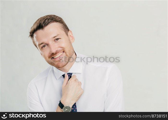 Cheerful unshaven male keeps hand on tie, dressed in elegant white shirt, smiles friendly, happy to achieve success in life, being prosperous and rich, isolated over white background. Man leader