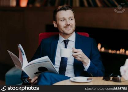 Cheerful unshaven male manager or employee has pleased expression, holds cup of coffee, rests in cafeteria, holds popular magazine, being focused aside, notices his colleague, dressed formally