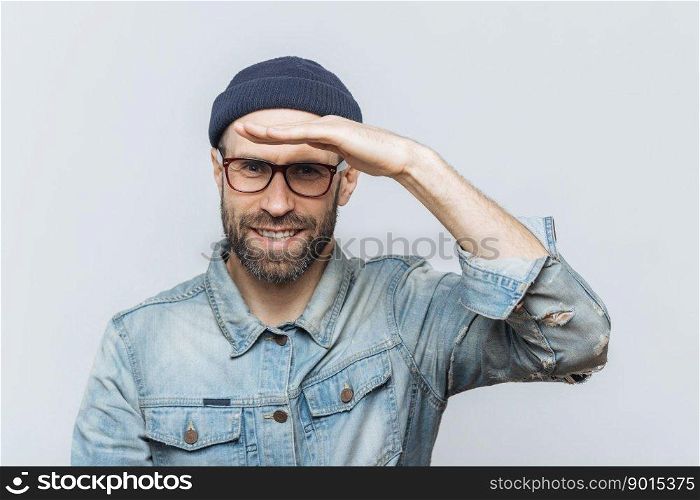 Cheerful unshaven middle aged male with thick stubble looks happily into distance, keeps hand near orehead, tries to see something, wears eyewear and denim jacket, isolated over grey background.