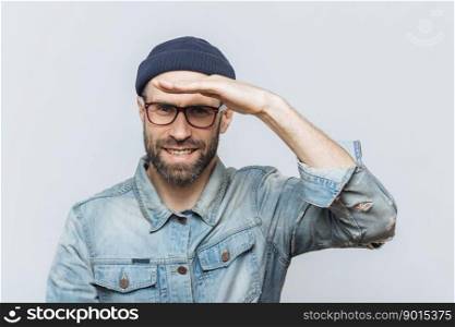 Cheerful unshaven middle aged male with thick stubble looks happily into distance, keeps hand near orehead, tries to see something, wears eyewear and denim jacket, isolated over grey background.