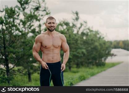 Cheerful unshaven sportsman skips with rope, has muscular body, enjoys fitness training, poses outdoor. Strong man with naked torso uses sport equipment for workout, breathes fresh air in nature