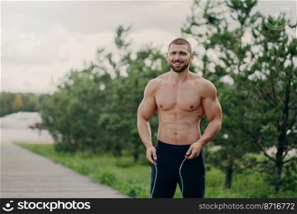 Cheerful unshaven sportsman skips with rope, has muscular body, enjoys fitness training, poses outdoor. Strong man with naked torso uses sport equipment for workout, breathes fresh air in nature