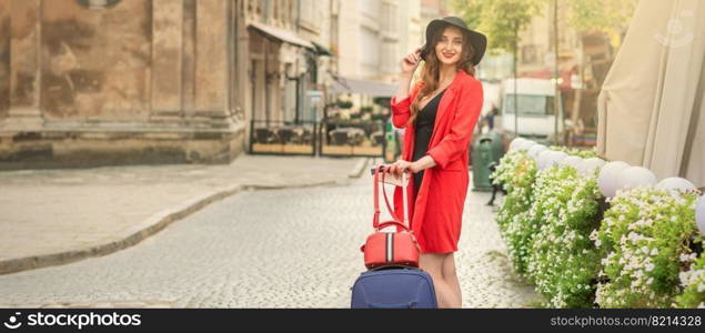 Cheerful white woman standing on the old street wearing hat with suitcase smiling at camera on background of city.. Close up portrait of fashionable young woman