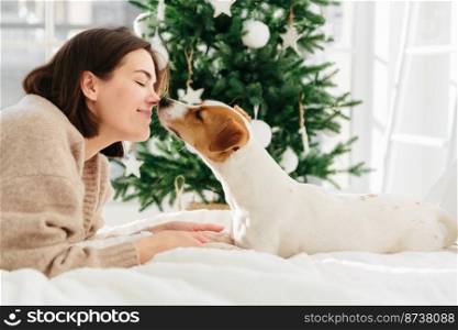 Cheerful woman gets kiss from favourite dog, keeps eyes closed from pleasure, lie on bed against Christmas tree at home, have nice relationship. People, New Year, celebration and pets concept