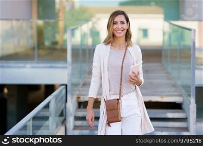 Cheerful woman in stylish clothes with smartphone smiling and looking at camera while standing on path near stairs outside modern building. Happy female with smartphone near steps