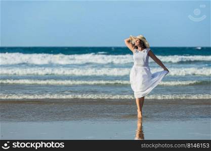 Cheerful woman in white dress touching straw hat and looking away with smile while walking on wet beach against sea waves and blue sky. Merry female tourist strolling near stormy sea