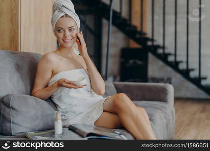 Cheerful woman of European appearance applies face cream for healthy soft skin, sits on comfortable sofa, surrounded by bottle of lotion and magazine, looks somewhere aside, has smooth comlexion