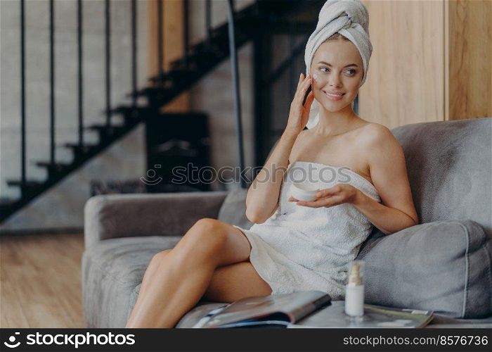 Cheerful woman of European appearance applies face cream for healthy soft skin, sits on comfortable sofa, surrounded by bottle of lotion and magazine, looks somewhere aside, has smooth comlexion