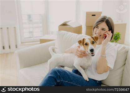 Cheerful woman poses on comfortable sofa, speaks with friend via smartphone, plays with pet, shares impression and good news about buying new flat, cardboard boxes in background. Delivery service