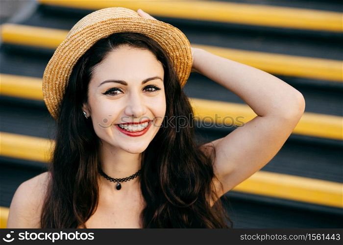 Cheerful woman with dark hair, healthy skin, bright eyes wearing straw hat on head sitting at stairs keeping her hand on head smiling broadly at camera. Carefree female resting alone. Emotions concept