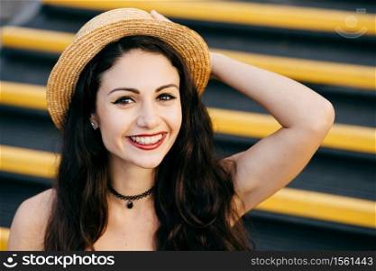 Cheerful woman with dark hair, healthy skin, bright eyes wearing straw hat on head sitting at stairs keeping her hand on head smiling broadly at camera. Carefree female resting alone. Emotions concept