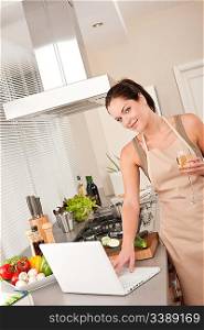 Cheerful woman with glass of white wine and laptop in the kitchen cooking