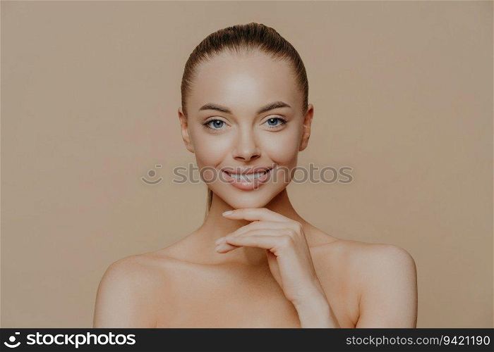 Cheerful woman with glowing skin, hand under chin, gentle smile, groomed body, combed hair. Pure skin, beauty and wellbeing.