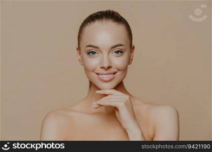 Cheerful woman with glowing skin, hand under chin, gentle smile, groomed body, combed hair. Pure skin, beauty and wellbeing.