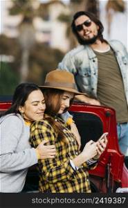 cheerful women embracing lady with smartphone near car trunk man leaning out from automobile