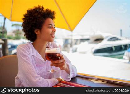 Cheerful young African American female in white blouse with curly hair smiling and looking away, while enjoying beer in street restaurant on sunny summer day on embankment. Happy black woman resting in street restaurant
