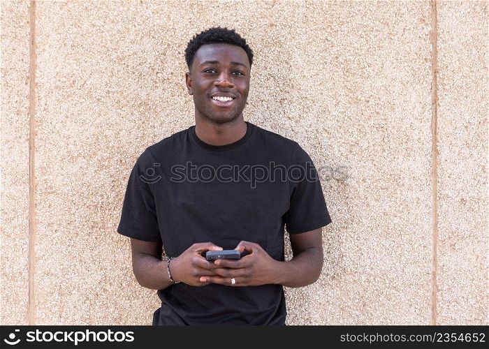 Cheerful young African American guy with dark curly hair in black t shirt smiling and looking at camera while messaging on smartphone against beige wall on street. Positive black man using smartphone and smiling at camera on street