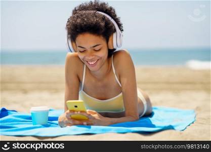 Cheerful young African American woman with curly dark hair, in bikini smiling and using mobile phone while, listening to music in headphones and lying on sandy beach on sunny day. Delighted ethnic female tourist in headphones browsing smartphone on seashore