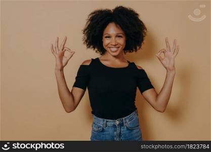 Cheerful young african female dressed in black tshirt shows ok sign okay gesture with both hands and demonstrates approval, isolated over beige background, smiling at camera and saying everything fine. Young african female shows ok sign okay gesture with both hands, isolated over beige background