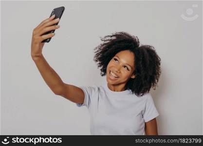 Cheerful young african woman with curly hair taking selfie on her modern mobile phone, looking at camera smiling happily while posing isolated over white studio background, wearing white basic tshirt. Cheerful young african woman with curly hair taking selfie on her modern mobile phone