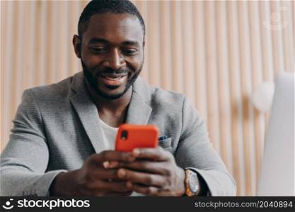 Cheerful young afro american man in stylish suit chatting online on smartphone looking on phone screen with joyful smile. African businessman enjoying correspondence with close friend or good customer. Cheerful young afro american man chatting online on smartphone looking on phone screen with smile