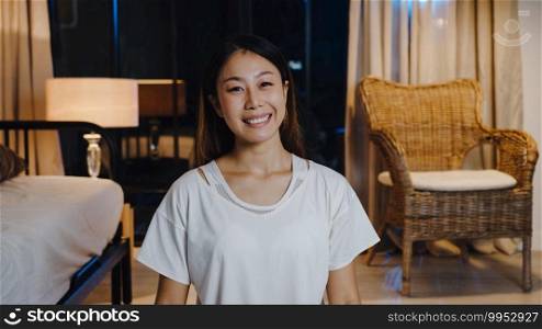 Cheerful young Asia lady feeling happy smile and look at camera using phone make live video call in living room at home night. Social distancing, quarantine for coronavirus. Close up webcam view.