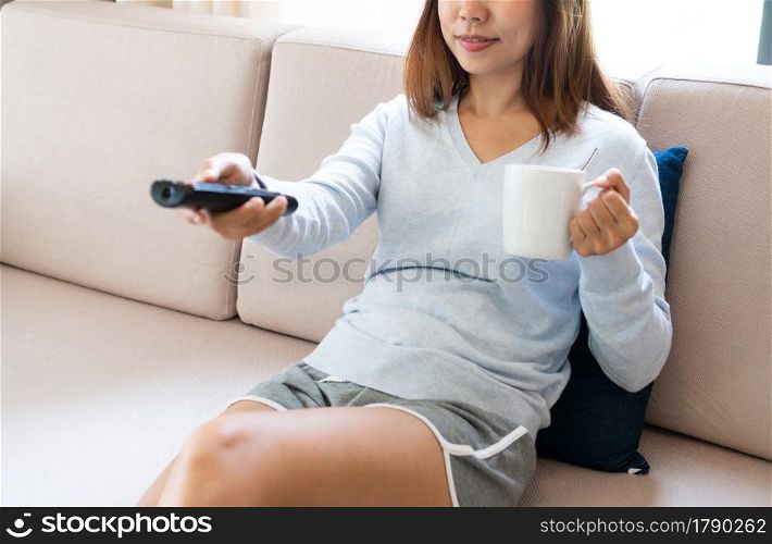 Cheerful young Asian woman relaxing, watching TV while drinking coffee on couch at home. Closeup