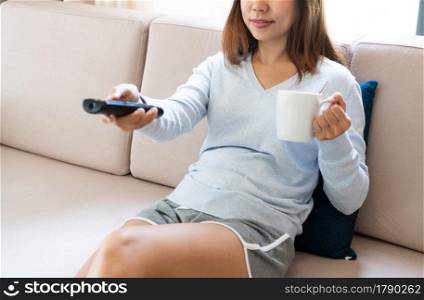 Cheerful young Asian woman relaxing, watching TV while drinking coffee on couch at home. Closeup
